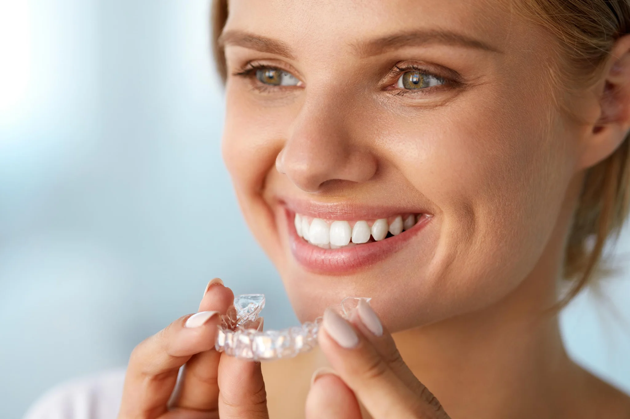 Woman smiling while holding clear dental aligner, demonstrating orthodontic treatment in bright clinical setting.